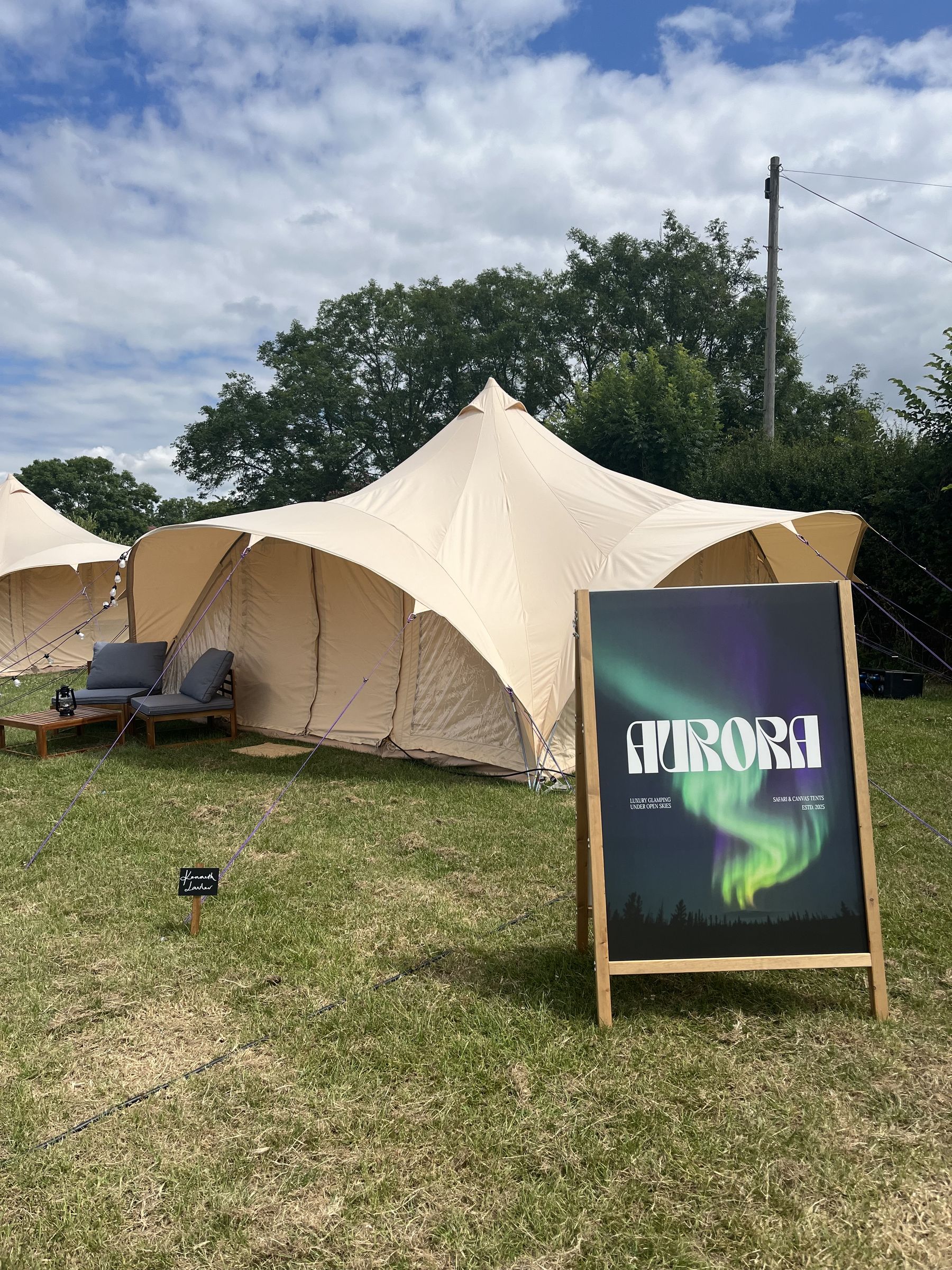 An Aurora A-board beside a pitched canopy, with a small named chalkboard staked in the grass